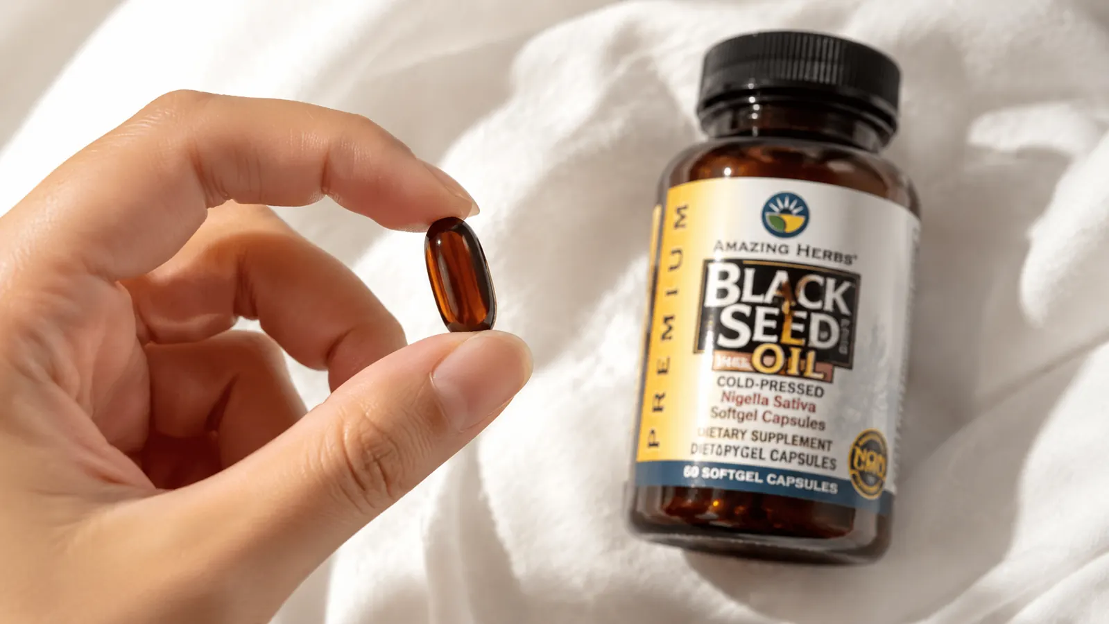Close-up of black seed oil softgel capsule held between thumb and forefinger, amber color visible through the shell